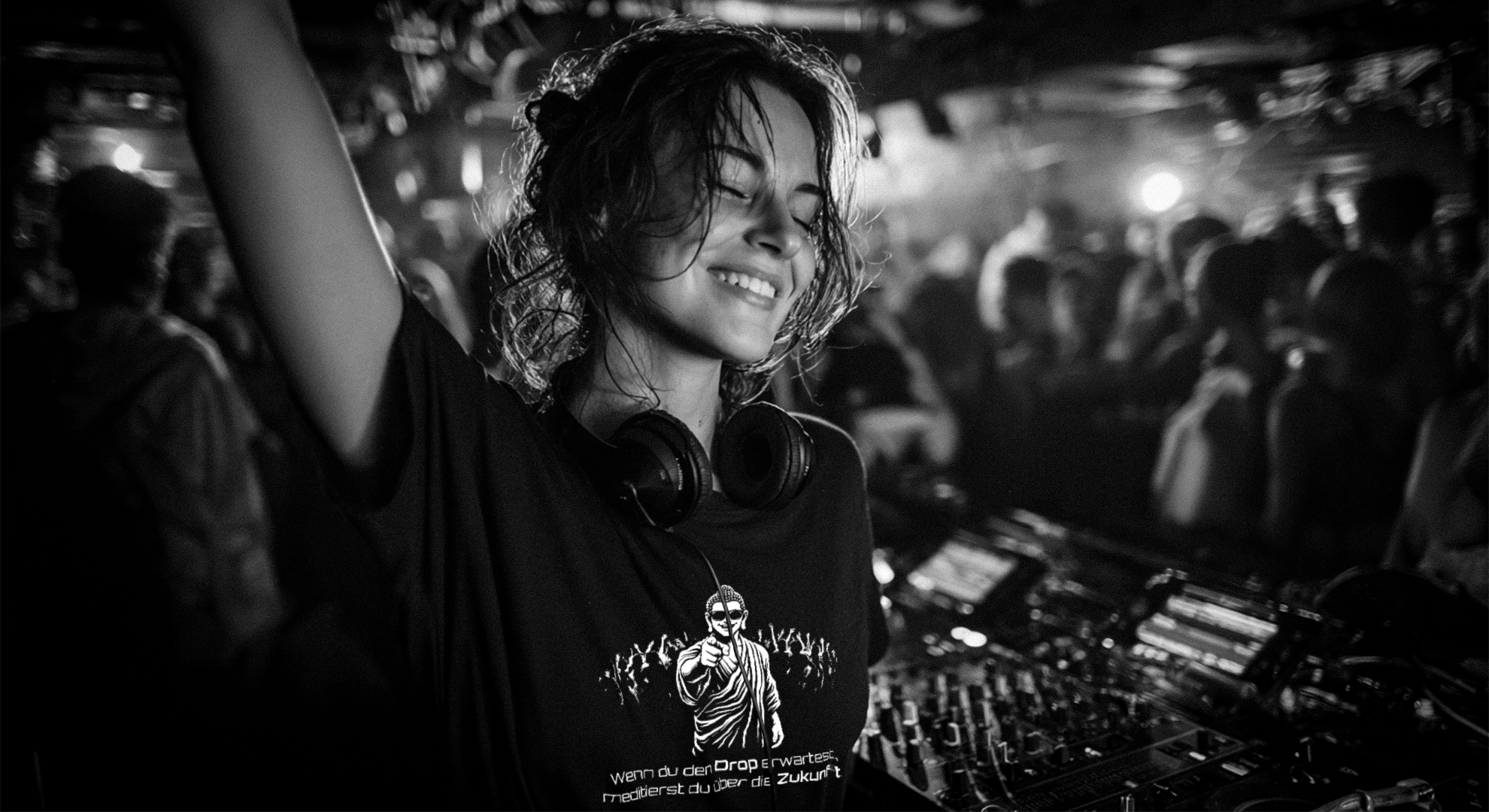 DJane auf Technoparty trägt schwarzes T-Shirt mit Buddha-Motiv und Spruch „Wenn du den Drop erwartest, meditierst du über die Zukunft“